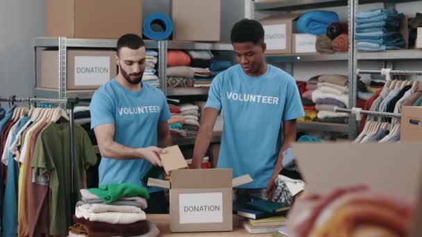 Δύο άντρες εθελοντές διπλώνουν βιβλία και τα βάζουν στο ντουλάπι της αποθήκης. Ανθρωπιστική βοήθεια και δωρεά ιδεών. Πλήρης προβολή μήκους. — Αρχείο Βίντεο
