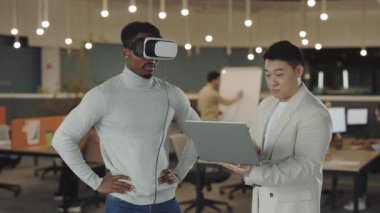Erkek arkadaşı verileri analiz edip bilgisayarda kayıt yaparken Afrikalı Amerikalı iş adamı sanal gerçeklik simülatörü kulaklığını test ediyor. Ofiste çalışanlar.
