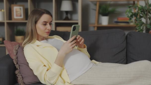 Akıllı telefonuyla kanepede yatan hamile beyaz kadın. — Stok video