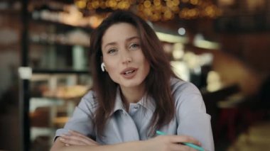 Kablosuz kulaklıklı kendine güvenen genç bir kadının portresi kafede oturuyor ve video görüşmesi sırasında kameraya konuşuyor. İnsan kavramı, röportaj ve vloglama.