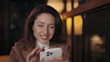 Pleasant caucasian woman in stylish jacket sitting at cozy cafe with modern smartphone in hands. Young lady smiling sincerely while looking on mobile screen.