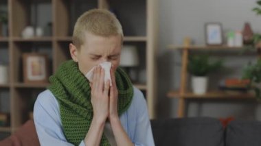 Kafkasyalı, kısa saçlı, peçeteye sümküren, kanepede boynunda sıcak eşarpla oturan hasta bir kadın. Hastalık, sağlık ve insan konsepti.