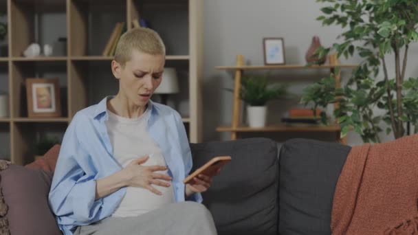 高加索女人期待着坐在沙发上 手里拿着智能手机 突然感到肚子痛 不舒服 健康状况和怀孕观念 — 图库视频影像