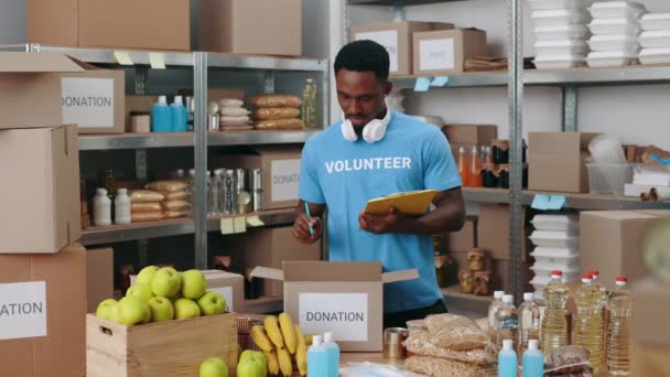 身穿蓝色志愿T恤的非裔美国人使用剪贴板在储物箱包装时做笔记 人与志愿工作的概念 — 图库视频影像