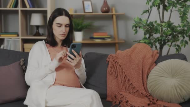 Вагітна жінка сидить на дивані з мобільним телефоном в руках — стокове відео