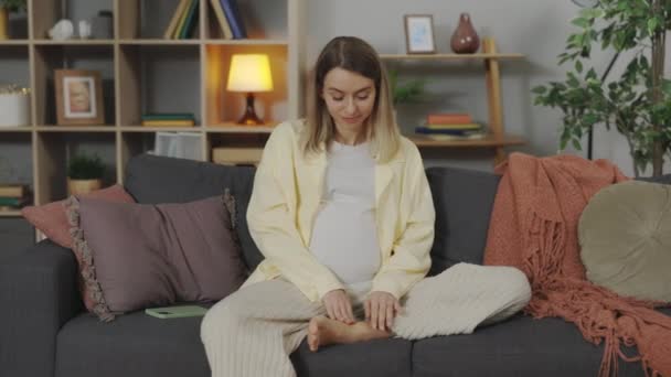 Pregnant woman sitting on sofa and massaging foot — Stock Video