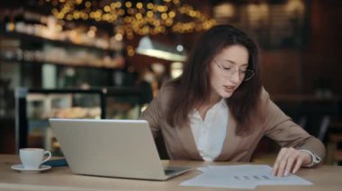 Kadın bazı belgeleri kontrol ediyor ve kafede dizüstü bilgisayar kullanıyor.