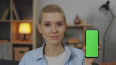 Smiling woman holding smartphone with green screen at home