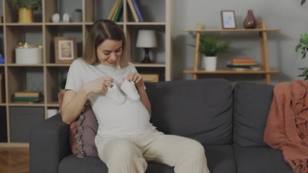 Young woman holding baby boots on pregnant tummy at home — Video