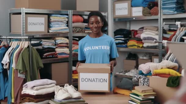 Vista retrato da mulher voluntária multirracial segurando caixa com roupas para doação e olhando para a câmera com sorriso. Prateleiras com pertences no fundo. Conceito de doação. — Vídeo de Stock