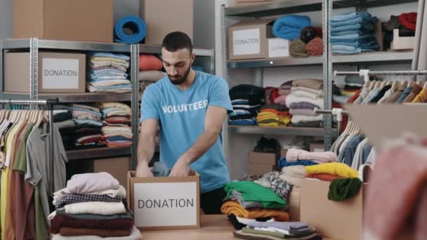Caucasian bearded male volunteer sorting clothes and putting it into the cupboard box at the warehouse. Humanitarian aid and donating concept. — Vídeo de stock