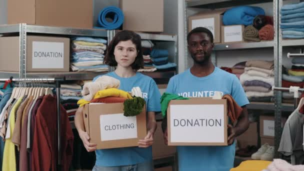 Πορτρέτο άποψη των διαφόρων εθελοντών κρατώντας κουτί ντουλάπα με λέξη δωρεά και κοιτάζοντας την κάμερα με χαμόγελο. Ράφια με αντικείμενα στο βάθος. Ανθρωπιστική βοήθεια. — Αρχείο Βίντεο