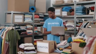 Çok ırklı erkek gönüllü, bağış merkezinde çalışırken dizüstü bilgisayar kullanıyor. Dolapta bir sürü kıyafet var. İnsani yardım ve gönüllüler konsepti.