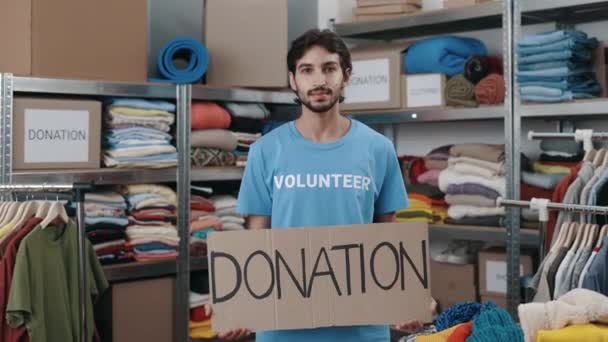 Portrait of energetic caucasian man looking at the camera and smiling while holding banner with donation word in charity shop. Volunteer working in organization and sorting donated clothes. — Stock Video