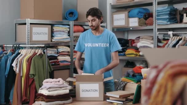 Voluntariado ordenando ropa donada en el centro de donación de caridad comunitaria. Trabajador masculino caucásico doblando ropa y poniéndola en la caja del armario en el almacén. Concepto de ayuda humanitaria. — Vídeo de stock