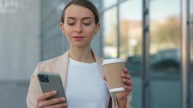 Elinde akıllı telefon ve kahve fincanı olan bir kadın ofisin yakınında yürüyor.