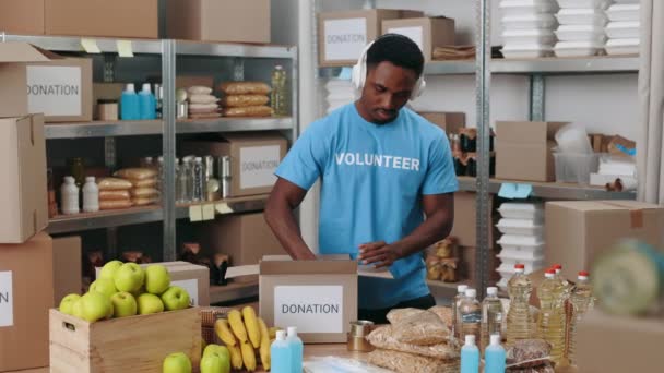 アフリカ系アメリカ人の男性が倉庫に立って 段ボール箱に缶詰を入れています 慈善財団で寄付を準備する男性ボランティア — ストック動画