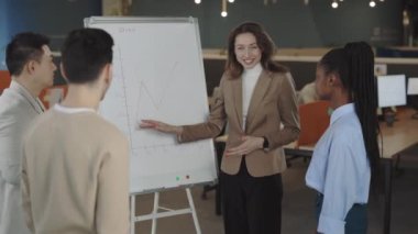 Olumlu iş kadını çok kültürlü personelin önünde duruyor ve reklam panosunda iyi satış sonuçları gösteriliyor. Koyu renk saçlı hoş bir bayan. Çalışanları planı yerine getirdikleri için tebrik ediyor..
