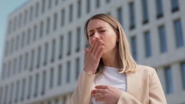 Ofis binasının yanında dikilen genç beyaz kadın öksürüyor. Sıradan şık giyinen çekici bir kadın hasta hissediyor ya da soğuk algınlığının ilk belirtilerini gösteriyor..