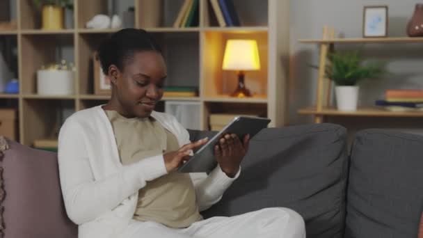 소파에 태블릿을 사용하여 복장을 아프리카 미국인 어머니를 미소짓고 기분좋은 온라인에서 — 비디오