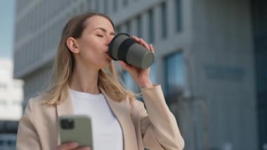 Genç, meşgul bir iş kadını akıllı telefon kullanıyor ve sokakta sıcak içeceklerin tadını çıkarıyor. Kahve molası sırasında ofis binasının yakınında gülümseyen güzel bir kadın..