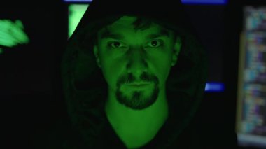 Arka planda yeşil bilgisayar görüntüleriyle karanlık bir sığınakta otururken kameraya bakan kendine güvenen beyaz hacker portresi. Siber güvenlik ve suç kavramı.