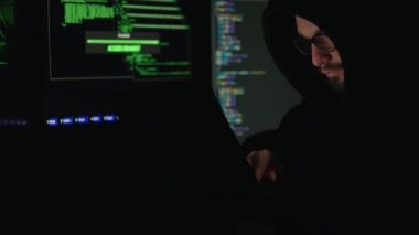 Gözlüklü beyaz sakallı bir adam çalışma yerinde oturuyor ve etrafında birden fazla dijital ekran var. Gizli organizasyon sistemine girmek için taşınabilir laptop klavyesinde şifreleri yazan kapüşonlu bilgisayar korsanı..