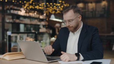 Yorgun işadamı mali belgeleri kontrol ederken kaşlarını çatıyor, koluyla alnına dokunuyor, dizüstü bilgisayarının modaya uygun kafesinde oturuyor, müşteriye internetten danışıyor, istatistikleri analiz ediyor, aşırı çalışıyor.