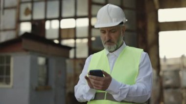 Yansıtıcı yelek ve beyaz miğfer giymiş, inşaat alanında beklerken cep telefonu kullanan olgun bir ustabaşı. Modern akıllı telefondan bina bilgilerini kontrol eden düşünceli bir adam..