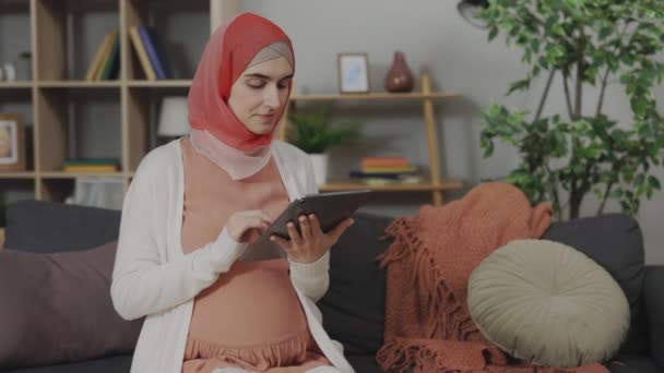 Mulher Grávida Árabe Usando Comprimido Digital Enquanto Sentado Sofá Confortável — Vídeo de Stock