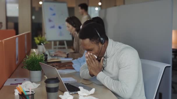 ノートパソコンで働いている間 ヘッドセットのくしゃみやナプキンの鼻を吹いている不健康なアフリカ系アメリカ人の男性従業員 背景に多様な同僚とオフィスでアレルギー症状を持つ病気の労働者 — ストック動画
