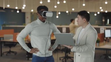 Erkek arkadaşı verileri analiz edip bilgisayarda kayıt yaparken Afrikalı Amerikalı iş adamı sanal gerçeklik simülatörü kulaklığını test ediyor. Ofiste çalışanlar