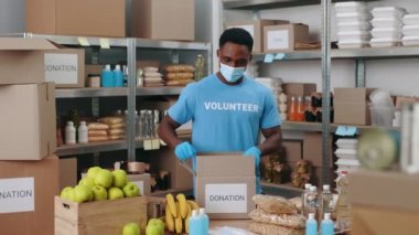 Afro-Amerikan maskeli ve eldivenli bir adam bağış için karton kutular ve yiyecek paketliyor. Depoda duran ve salgın sırasında teslimat için paketleri hazırlayan gönüllü..