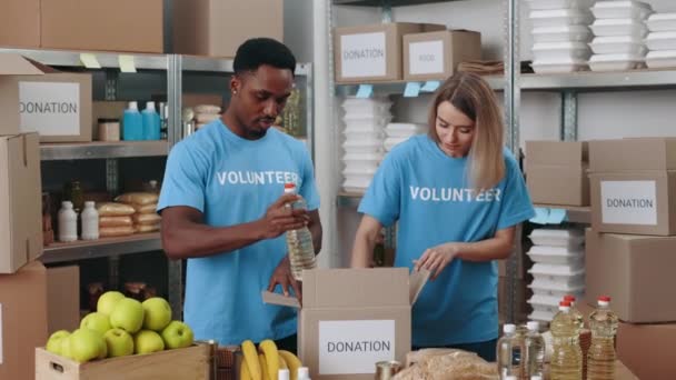 多人種の男性と女性のボランティアが一緒に倉庫に立っている間 段ボールの寄付箱の中に食べ物を入れます チームワーク 人道の概念 — ストック動画