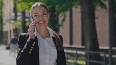 Şık resmi giyinen neşeli bayan girişimci ofis merkezinin yanındaki modern akıllı telefondan konuşuyor. Beyaz kadın dışarıda dolaşırken iş haberlerini tartışıyor..