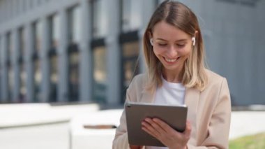Kablosuz kulaklıkla gülümseyen iş kadını dijital tablet kullanıyor ve modern ofis merkezinin yanında dururken sosyal medyayı kontrol ediyor. İnsan kavramı, yaşam tarzı ve aletler.