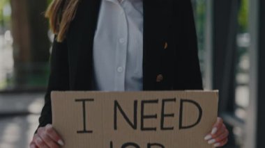 Şık giyinmiş, elinde karton pankartla şehir merkezinde dururken işe ihtiyacım olduğunu söyleyen genç bir kadın. İnsan kavramı, işsizlik krizi ve kariyer kaybı.