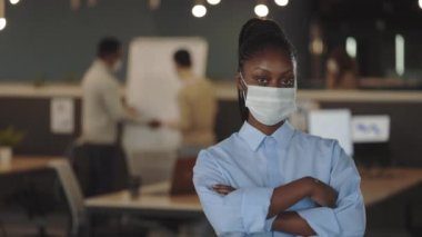 Tıbbi maske takmış genç Afro-Amerikan iş kadını modern ofiste duran kameraya bakıyor. Ofis odasındaki bir adamın portresi. Coronavirus konsepti