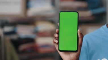 Çok ırklı bir kadın elinde yeşil renkli akıllı telefon tutuyor ve depoda dikilirken kameraya gösteriyor. Gönüllü konsepti. Kırpılmış görünüm