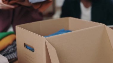 Kafkasyalı bir kadın, meslektaşı ile tekrar kullanım merkezinde çalışırken kıyafetlerini sınıflandırıyor ve kutuya koyuyor. İkinci el ve insani yardım kavramı. Kırpılmış görünüm