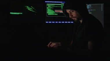 Geceleri karanlık odada modern dizüstü bilgisayarda ciddi genç hacker programları. Siyah kapüşonlu konsantre genç adam virüslü yazılıma saldırmak için bilgisayar bilgisini kullanıyor..