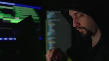 Siyah kapüşonlu sakallı erkek hacker elinde modern taşınabilir bilgisayarla karanlık odada oturuyor. İnternet teröristleri sisteme tehlikeli siber virüs bulaştırıyor..