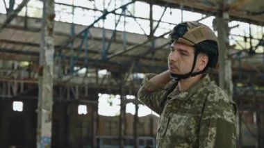 Kafkaslı ve kamuflaj elbiseli beyaz adam terk edilmiş çelik fabrikasında yürüyor ve sırtında sırt çantası taşıyor. Görevdeki cesur asker. Ordu konsepti.