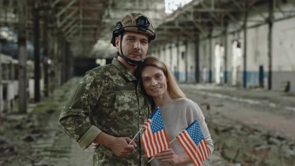 身穿军服的男战士站在被毁的工厂时拥抱可爱的妻子的画像 年轻的家庭微笑着看着相机 手里拿着美国国旗 — 图库视频影像