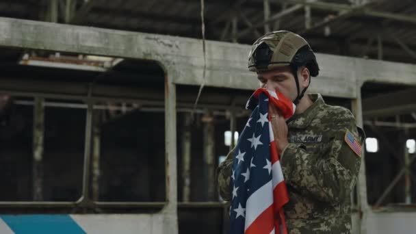 任務後の破壊された建物の中に立っている間 米軍の制服を着た男性兵士がアメリカ国旗にキス 武力の概念と国の誇り — ストック動画