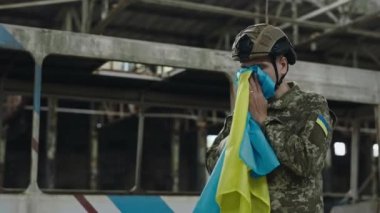 Kamuflaj üniforması ve şapkası içinde Ukrayna bayrağını sevgi ve dualarla tutan yorgun duygusal asker. Yok edilen fabrikanın bulanık arka planı. Askeri çatışma ve savaş kavramı.