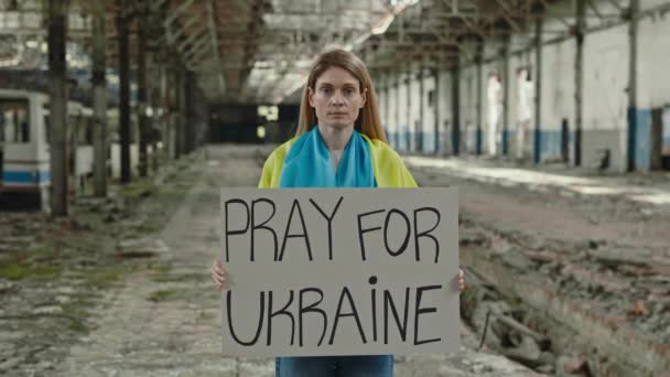 国旗を身に着けている悲しい女性の肖像画と台無し工場でウクライナのフレーズのための祈りとポスターを保持 ロシアの侵攻に抗議しながらカメラを見ている白人女性 — ストック動画