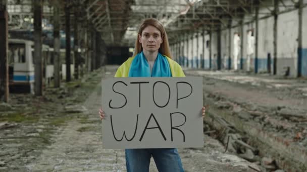 ストップ戦争のテキストと段ボールバナーを保持し 廃墟工場を背景にカメラを見て愛国的な白人女性の肖像画 ウクライナの平和を求める — ストック動画