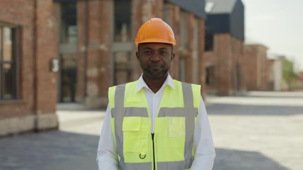 仕事中のアフリカ系アメリカ人男性が腕を組んで立ってカメラを見て笑っている姿が描かれている 建設現場で働く正のミドルビルダーの男 — ストック動画