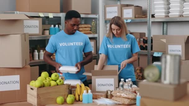 白人女性とアフリカ系アメリカ人男性が一緒に倉庫や募金箱に立っています フードバンクでボランティアをしている多人種間の人々 — ストック動画